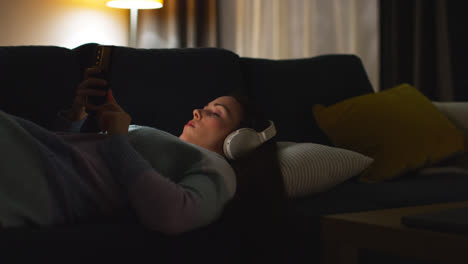 Mujer-Con-Auriculares-Inalámbricos-Recostada-En-Un-Sofá-En-Casa-Por-La-Noche-Transmitiendo-O-Mirando-Contenido-En-Línea-En-Un-Teléfono-Móvil-3